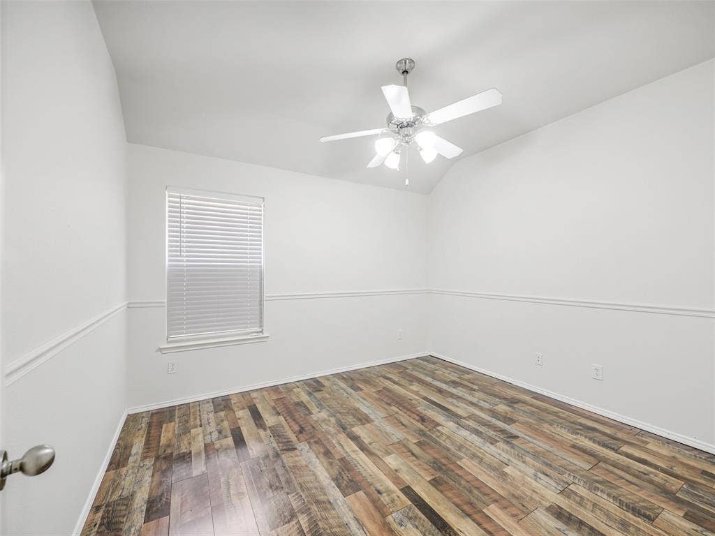 414 Dartmoor Drive, Celina, TX 75009 A spacious secondary bedroom at 414 Dartmoor Dr, Celina, TX. The room includes multi-tone wood-look plank flooring, detailed white chair rail trim along neutral walls, a white 5-blade ceiling fan with light kit, and a large window with 2-inch faux wood blinds for natural light.
