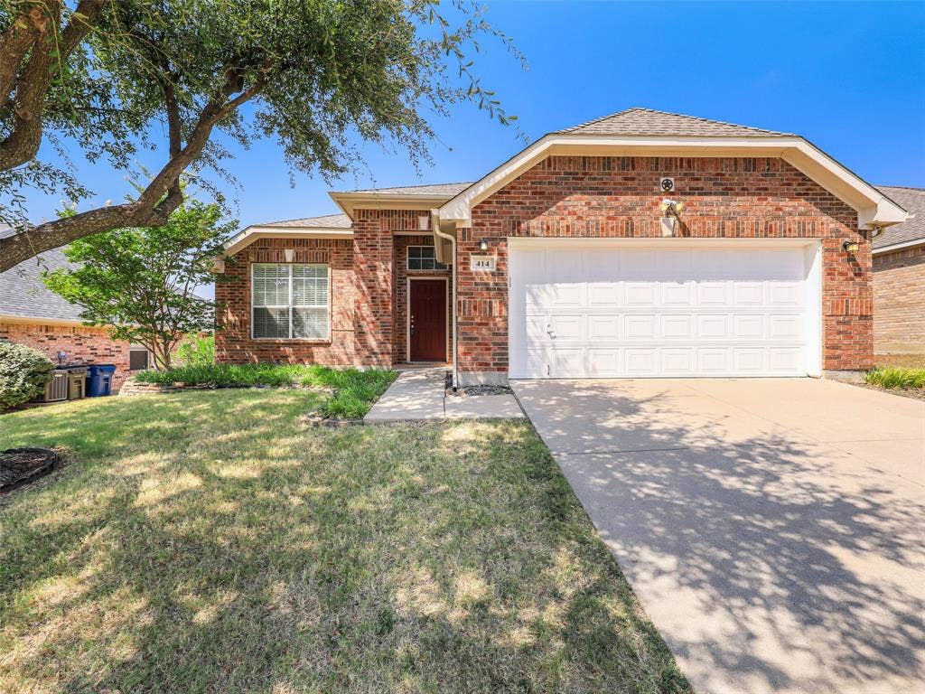 414 Dartmoor Drive, Celina, TX 75009 A close-up view of the front elevation at 414 Dartmoor Dr, Celina, TX 75009. The home, located in the Carter Ranch neighborhood, features classic red textured brick construction, a front-facing two-car garage with a white panel door, and a covered front porch entry with a red front door. The landscaped beds include native Texas grasses beneath mature shade trees.