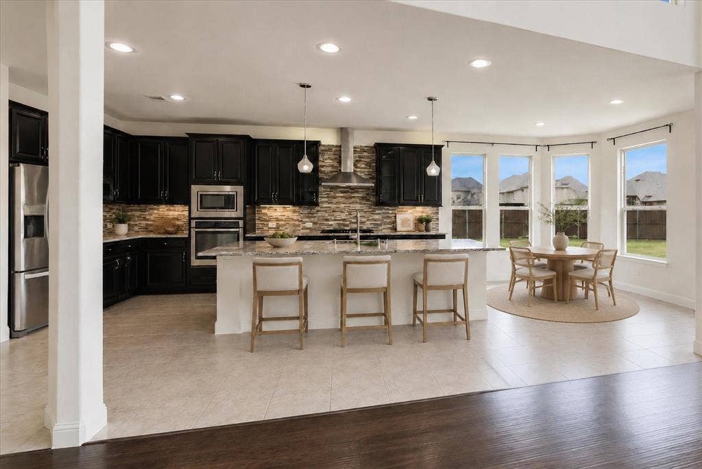 1650 Chisholm Trail, Prosper, TX 75078 Modern kitchen featuring dark cabinetry, stainless steel appliances, a stone-finish backsplash, a kitchen island with seating, and a dining area with bay windows