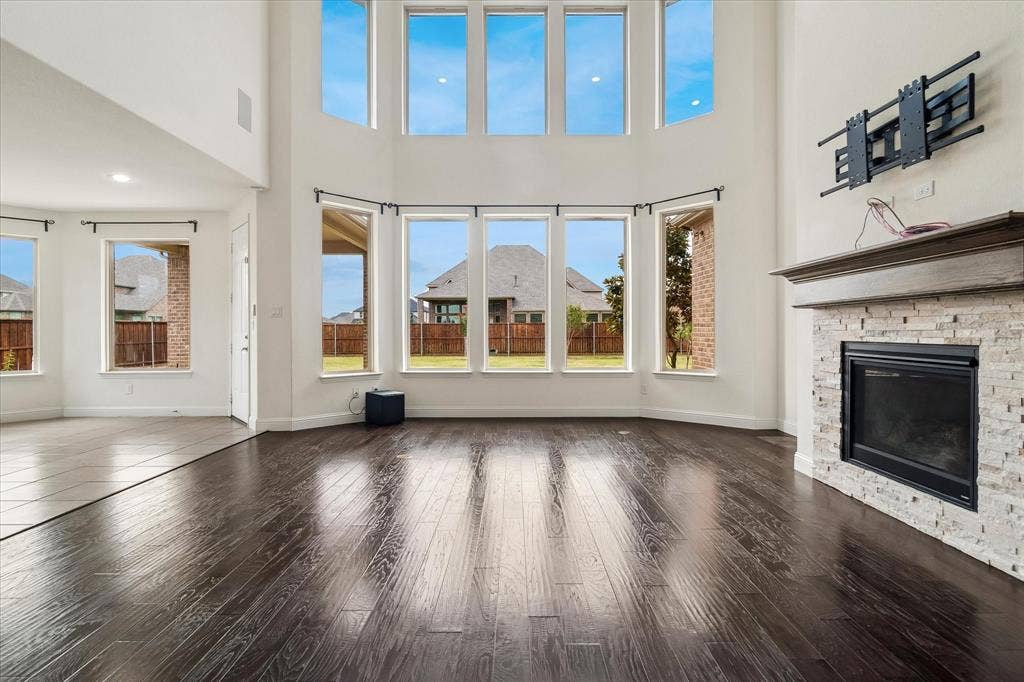 1650 Chisholm Trail, Prosper, TX 75078 Two-story living area featuring dark wood-finish flooring, a stone-faced fireplace with a wooden mantel, and a wall of windows offering natural illumination and yard views