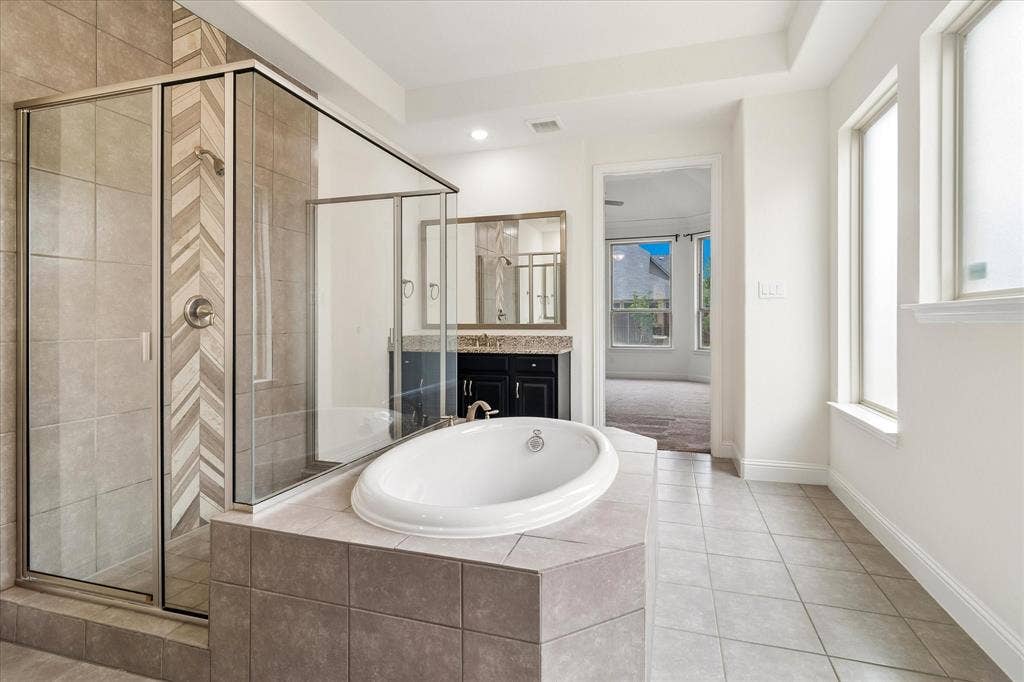 1650 Chisholm Trail, Prosper, TX 75078 Bathroom featuring a glass-enclosed shower with chevron-patterned tile accents, a built-in soaking tub with a tiled surround, a dark wood-finish vanity with a light-toned countertop, and multiple windows