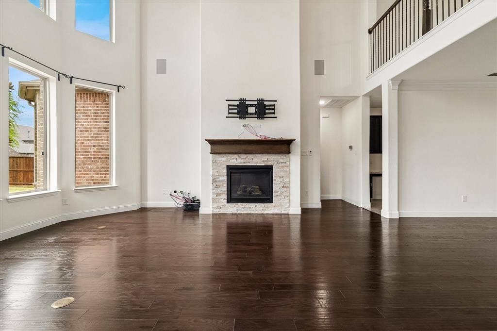 1650 Chisholm Trail, Prosper, TX 75078 Two-story living area featuring dark wood-finish flooring, a stone-clad fireplace with a wood mantel, high ceilings, multiple windows, and an upper-level railing