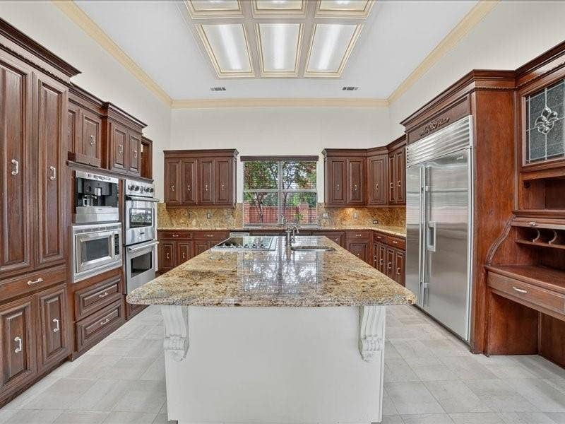 5201 Windjammer Road, Plano, TX 75093 Gourmet kitchen featuring a large central island with a cooktop, granite countertops, built-in stainless steel appliances, extensive dark wood cabinetry, and a coffered ceiling with integrated lighting