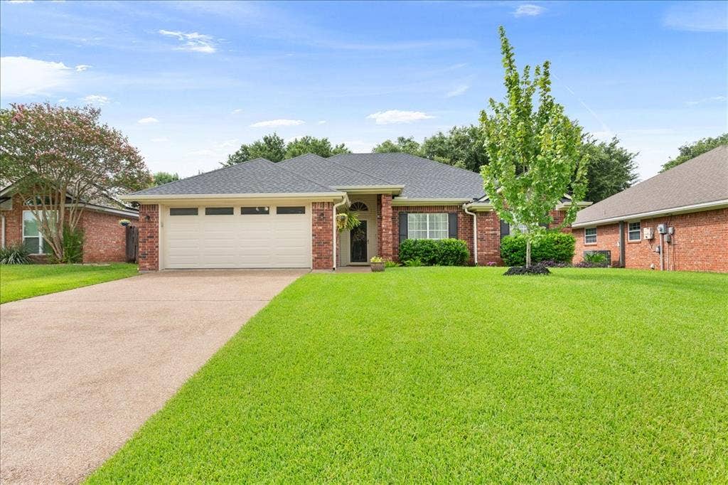 10308 Eagle Mountain Drive, Waco, TX 76712 Brick exterior featuring a two-car garage with transom windows, a dark shingle roof, a concrete driveway, and a verdant lawn with mature landscaping