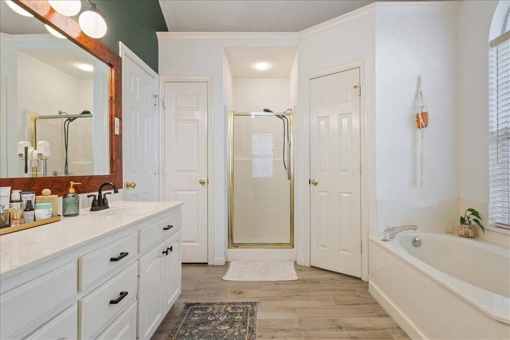 10308 Eagle Mountain Drive, Waco, TX 76712 Bathroom featuring a white vanity with black hardware, a wood-framed mirror, a built-in bathtub, a glass-enclosed shower, and wood-finish flooring