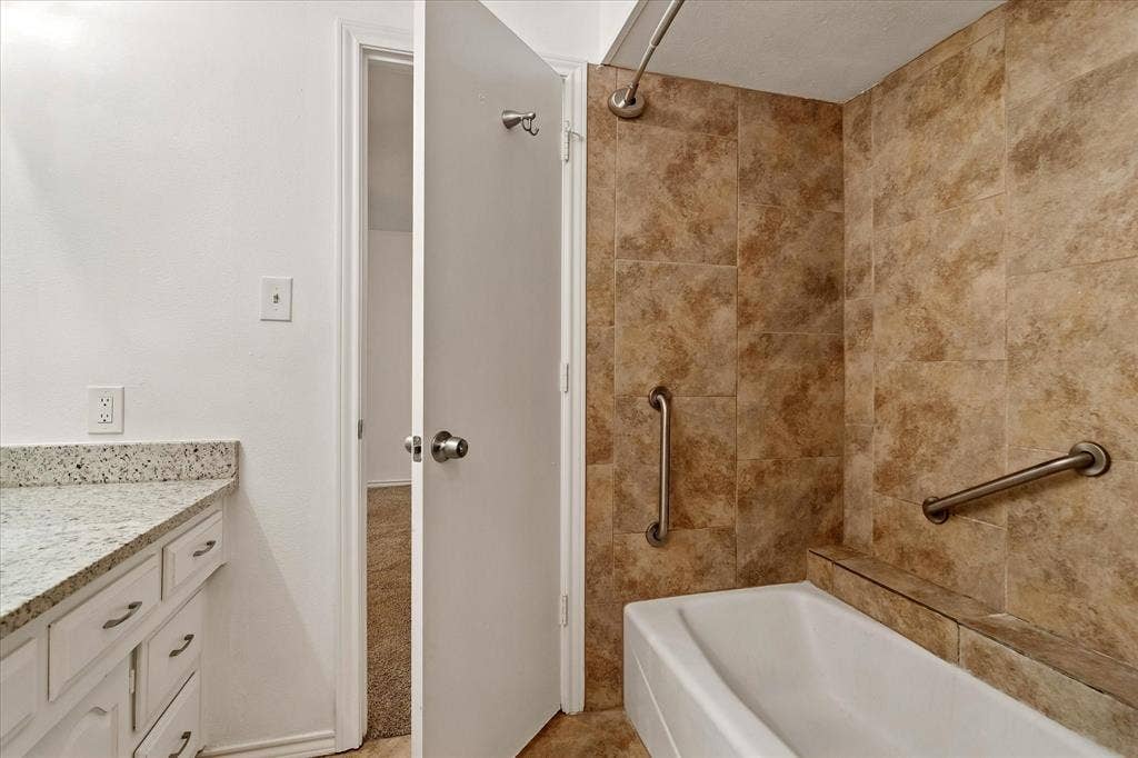 3318 Wells Drive, Plano, TX 75093 Bathroom featuring a white bathtub with a shower, brown tile surround, two grab bars, and a white vanity with a light-colored speckled countertop
