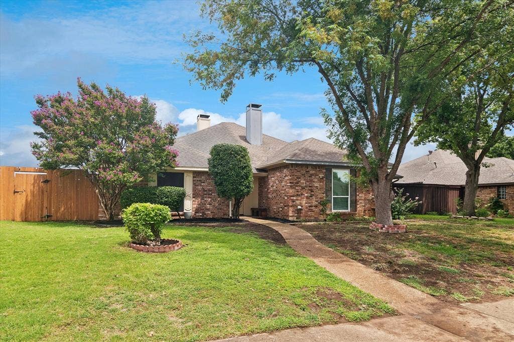 3318 Wells Drive, Plano, TX 75093 Traditional brick exterior with an attached garage, a mature tree canopy, a manicured lawn, a concrete walkway, and a privacy fence