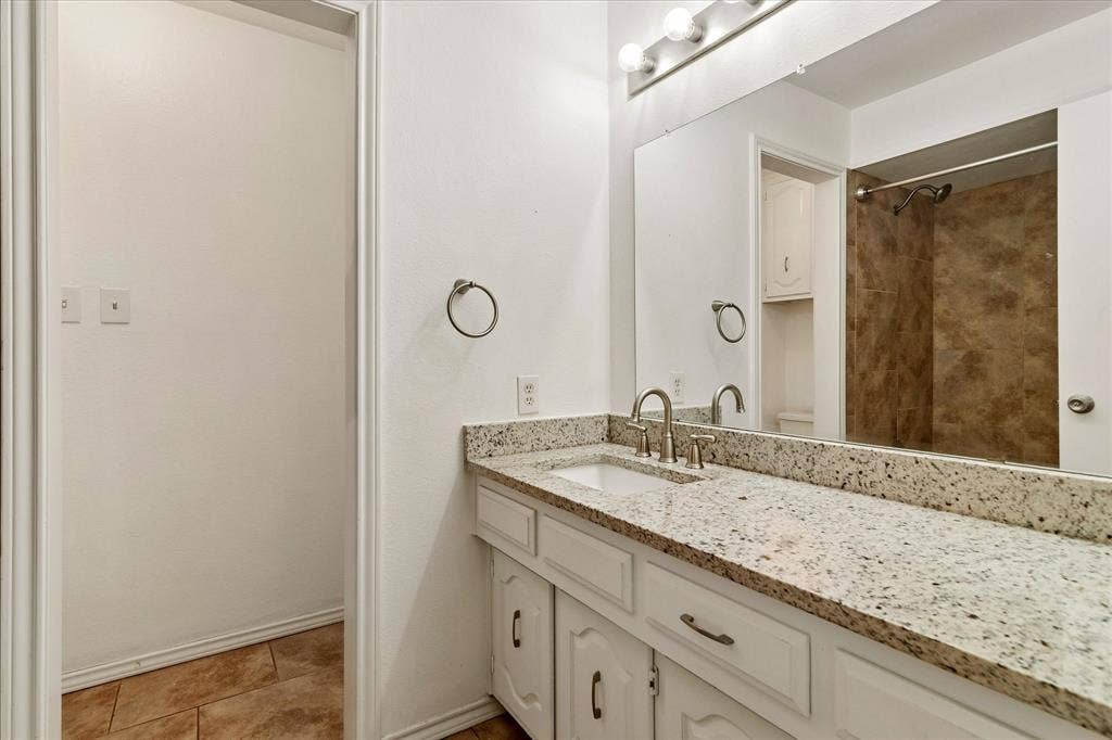 3318 Wells Drive, Plano, TX 75093 Bathroom featuring a granite-finish countertop, dual-handle faucet, white cabinetry with silver-tone hardware, and a mirrored vanity