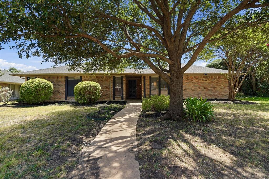1000 Lexington Drive, Plano, TX 75075 Brick exterior residence with a gray shingle roof, featuring a concrete pathway and mature trees providing shade