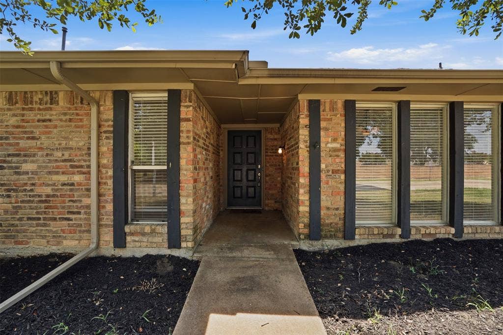 1000 Lexington Drive, Plano, TX 75075 Brick facade residence featuring a covered entry, dark paneled front door, and multiple front-facing windows with blinds