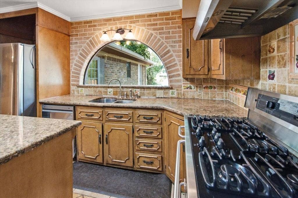 612 W Highland Street, West, TX 76691 Kitchen featuring an arched window with brick surround, wood-finish cabinetry, stainless steel appliances, gas range with vent hood, granite countertops, and decorative tile backsplash