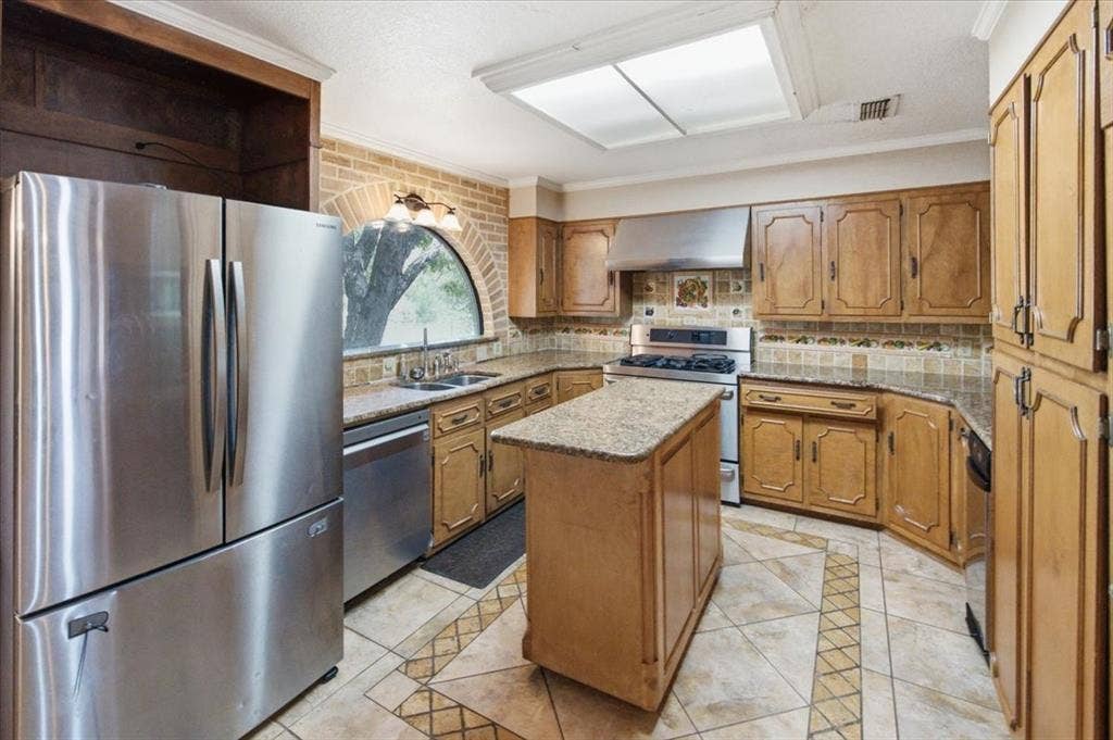 612 W Highland Street, West, TX 76691 Kitchen featuring an brick-arched window, wood-finish cabinetry, tile flooring with decorative inlay, stainless steel appliances, and a central island with a granite countertop