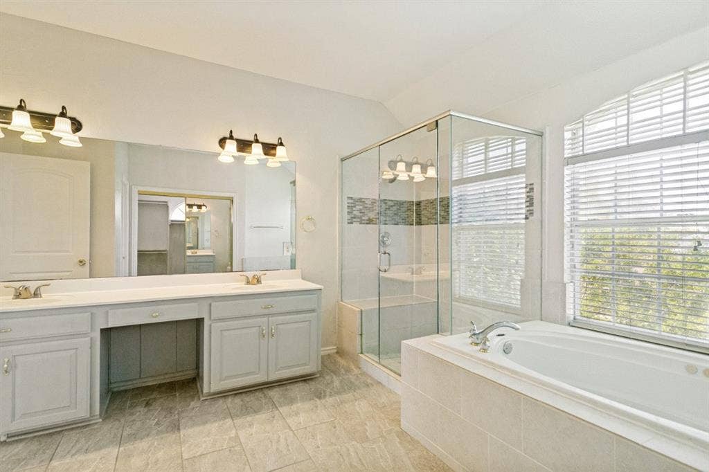 8512 Mesa Verde Drive, Plano, TX 75025 Expansive bathroom featuring a tiled floor, dual vanity with light cabinetry, large mirror, glass-enclosed shower with tiled accents, and a built-in soaking tub beneath a window