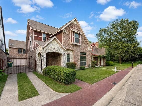 Stone and brick facade residence featuring an arched stone entry, a gated driveway leading to a garage, a red paver sidewalk, and a manicured lawn