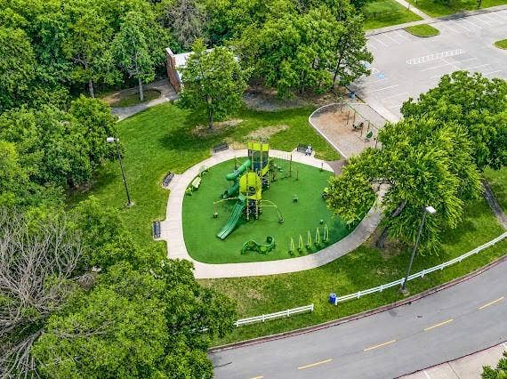 Expansive park area featuring a modern playground with artificial turf surfacing, dedicated swing set area, multiple seating benches, mature trees, and a paved walking path