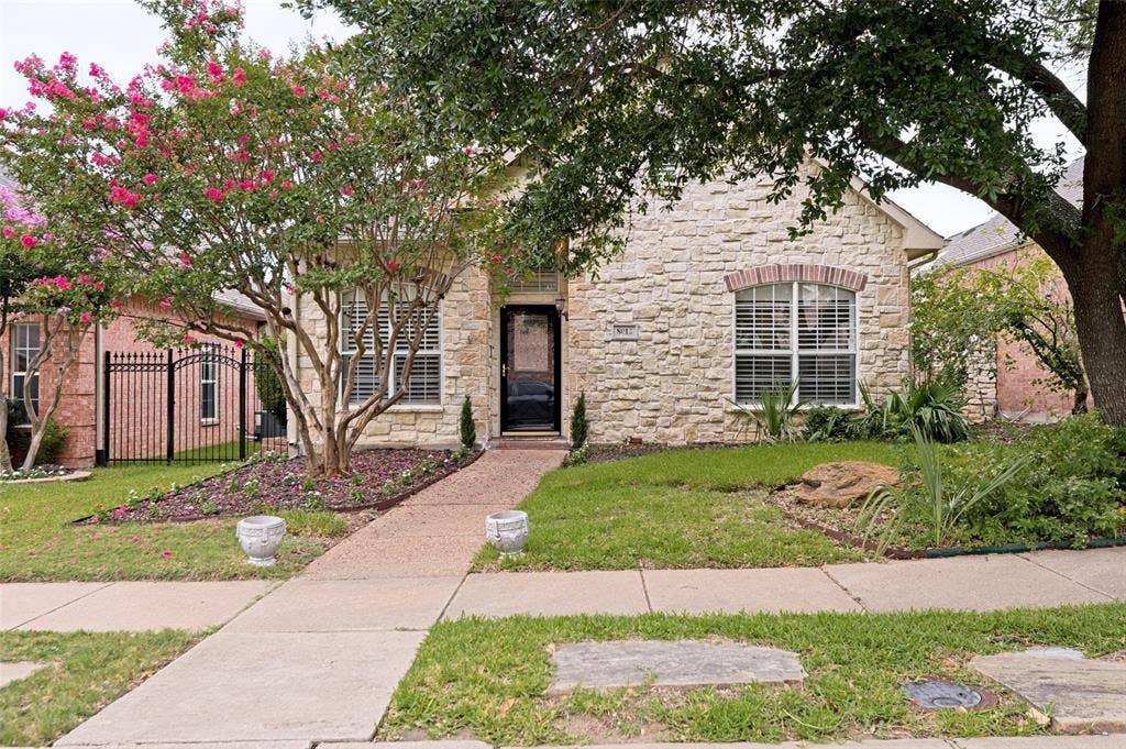 8012 Springmoss Drive, Plano, TX 75025 Stone facade residence featuring a dark paneled entry door, arched windows with shutters, and mature landscaping with flowering trees