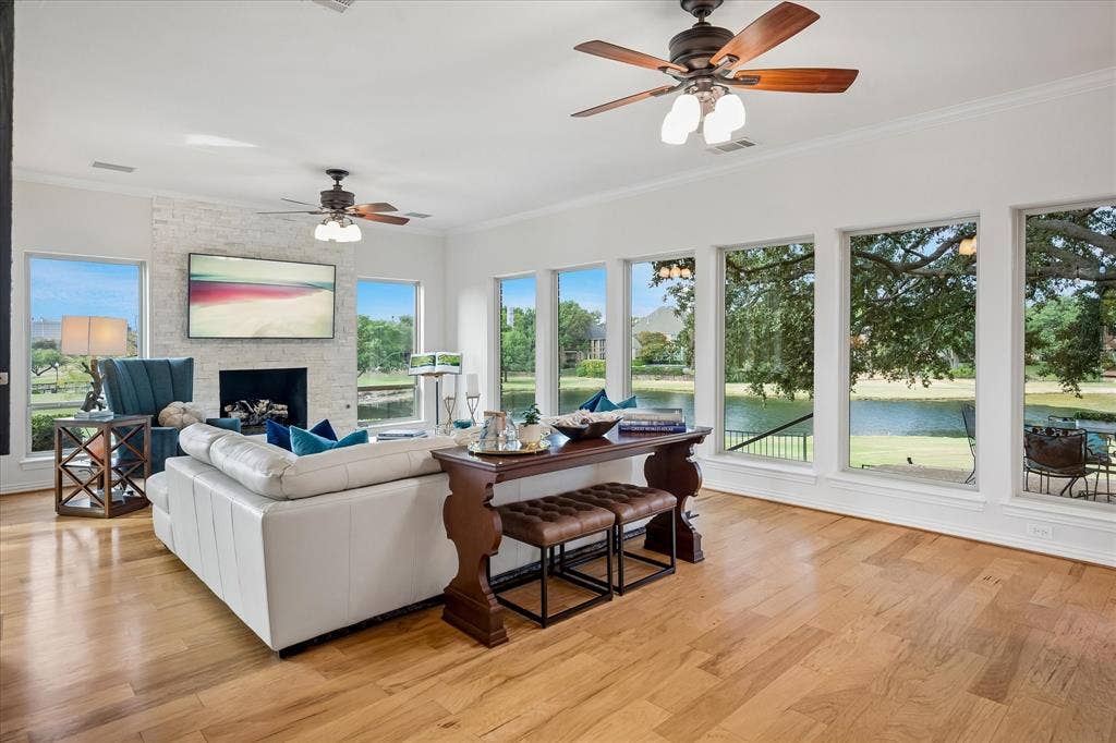 7501 Sweetgum Drive, Irving, TX 75063 Hardwood flooring, white brick fireplace, multiple ceiling fans, and large windows with water and tree scenery