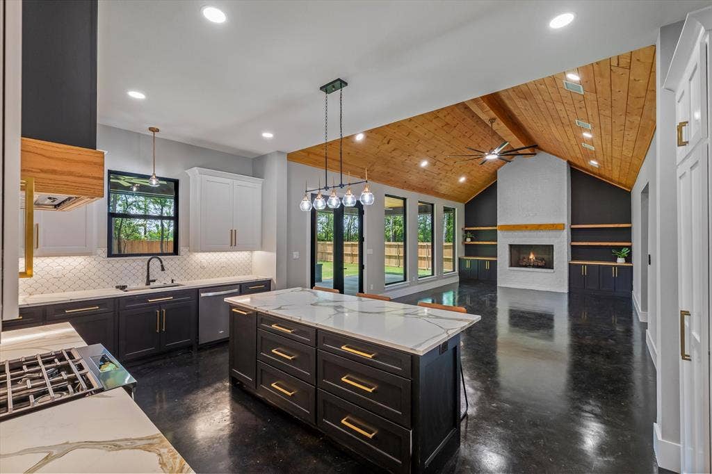 12253 Madison Xing, Bullard, TX 75757 Open-concept living space featuring a vaulted wood-paneled ceiling, a central kitchen island with a quartz countertop, a gas range with a modern vent hood, a white brick fireplace with a wood mantel, and built-in shelving and cabinetry