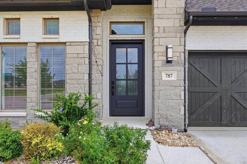 787 Cedarwood Court, Haslet, TX 76052 Modern exterior featuring a dark paneled entry door with glass inserts, light-toned stone and brick facade, a contemporary outdoor light fixture, and a dark wood-finish garage door