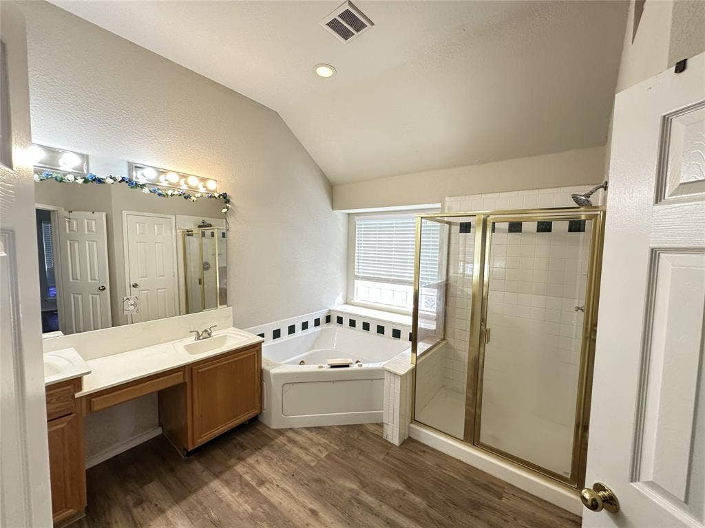 Spacious bathroom featuring wood-finish flooring, a dual-sink vanity with ample counter space, a corner soaking tub, and a separate shower enclosure