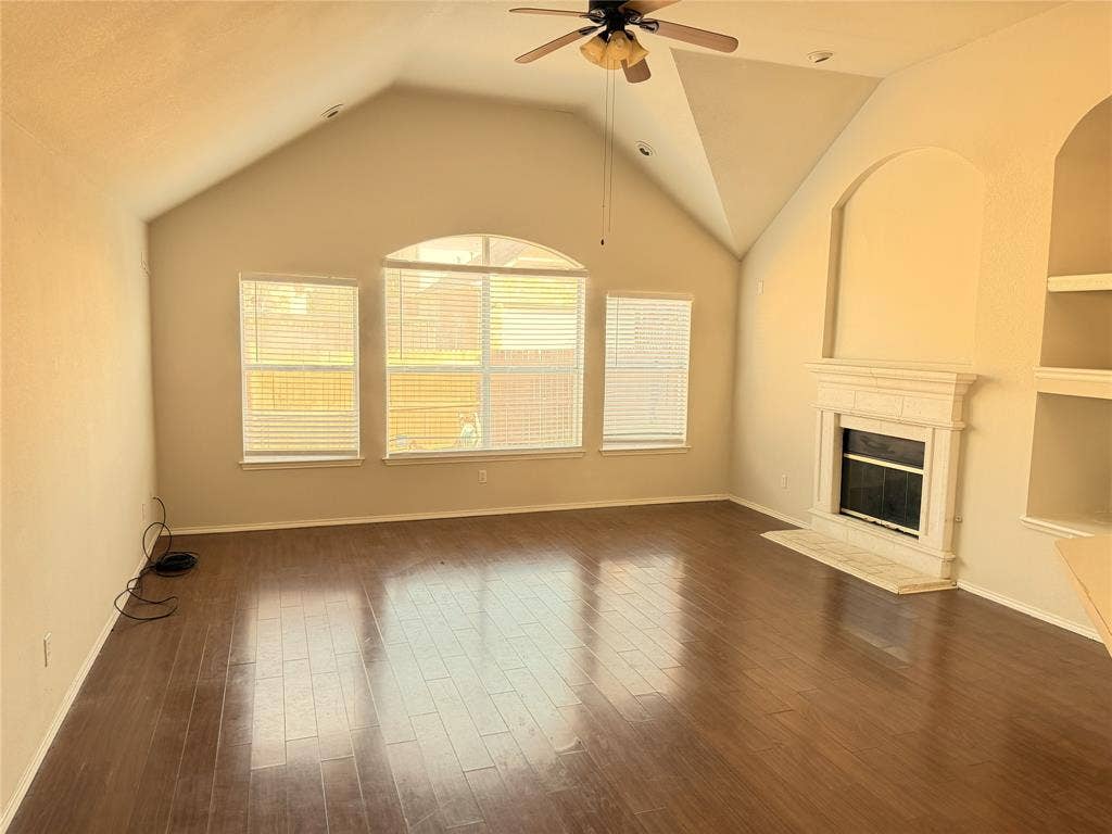 Vaulted ceiling with ceiling fan, wood-finish flooring, multiple windows with blinds, built-in fireplace with hearth, and built-in shelving