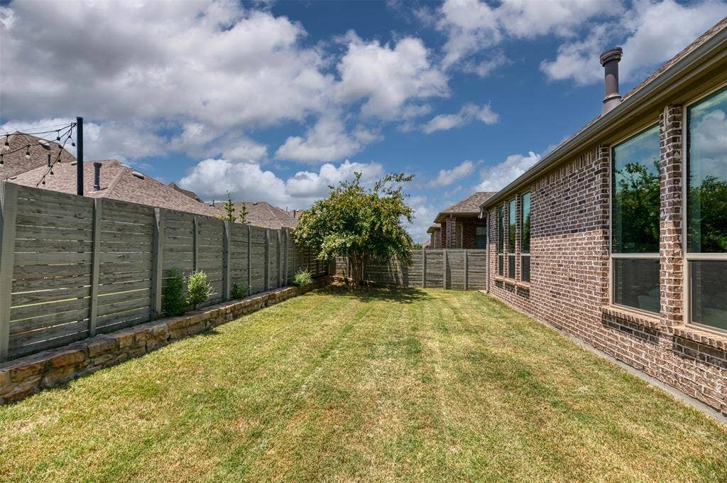 1109 10th Street, Argyle, TX 76226 Expansive grassy yard featuring a brick-clad exterior with multiple windows, a horizontal slat fence, a stone retaining wall, and mature landscaping