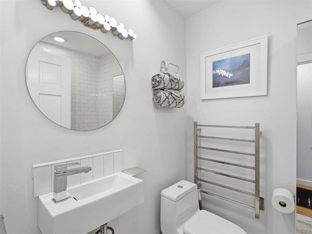 Contemporary bathroom featuring a wall-mounted rectangular sink with a single-handle chrome faucet, a circular mirror, a decorative vanity light fixture, a toilet, and a wall-mounted towel warmer