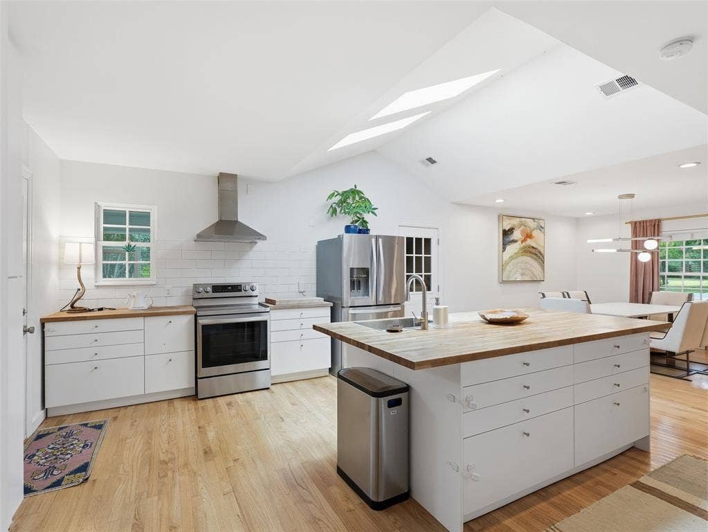 Vaulted ceilings with skylights illuminate this kitchen featuring stainless steel appliances, a center island with a wood-finish countertop, white cabinetry with silver hardware, and wood-finish flooring