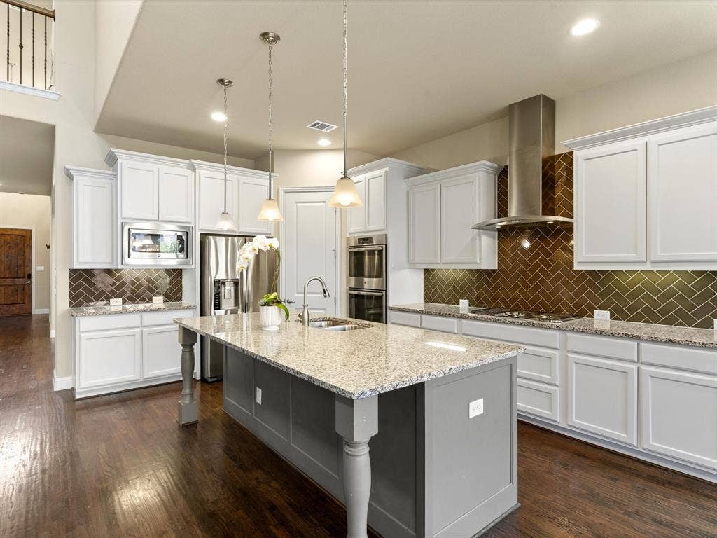 2703 Navarro Trail, Euless, TX 76039 Spacious kitchen featuring an expansive center island with a sink, stainless steel appliances, a herringbone tile backsplash, and dark wood-finish flooring