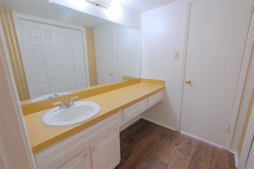 6317 Granbury Road, #6321, Fort Worth, TX 76133 Vanity area featuring a single basin sink, a large mirror, a laminate countertop, white cabinetry with storage, and wood-finish flooring