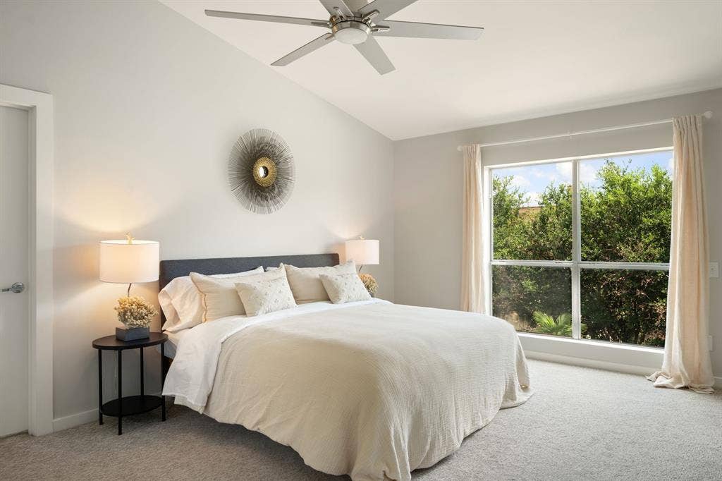 Vaulted ceiling with a ceiling fan, broad window with outdoor greenery, light-toned carpet flooring, white door with brushed nickel hardware, and neutral wall tones