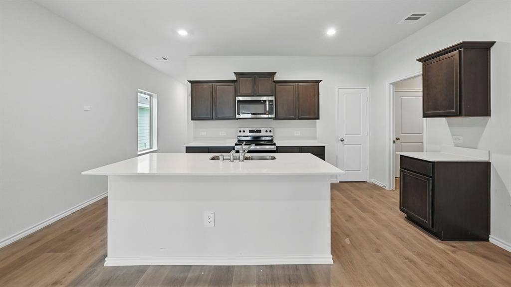 5602 Gannet Place, Crandall, TX 75114 Modern kitchen featuring an expansive island with an integrated sink, dark wood-finish cabinetry, light countertops, stainless steel appliances, and wood-finish flooring
