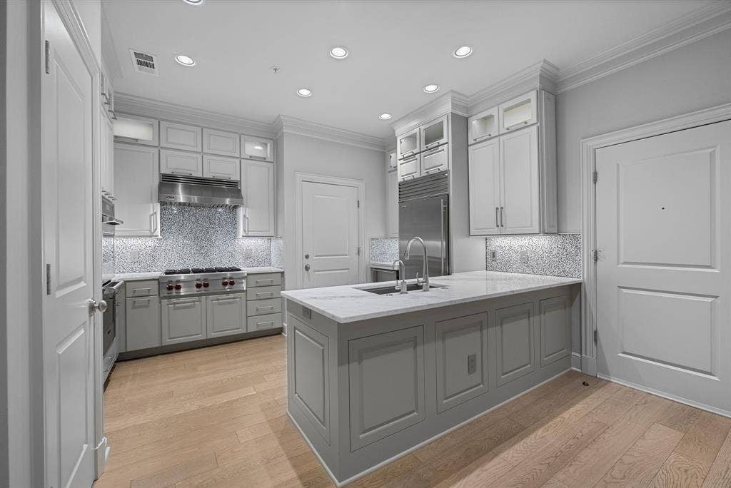 4240 Prescott Avenue, #8B, Dallas, TX 75219 Gourmet kitchen featuring a large central island with an undermount sink, a professional-grade gas range with stainless steel hood, custom cabinetry with glass-front uppers, a mosaic tile backsplash, and wood-finish flooring
