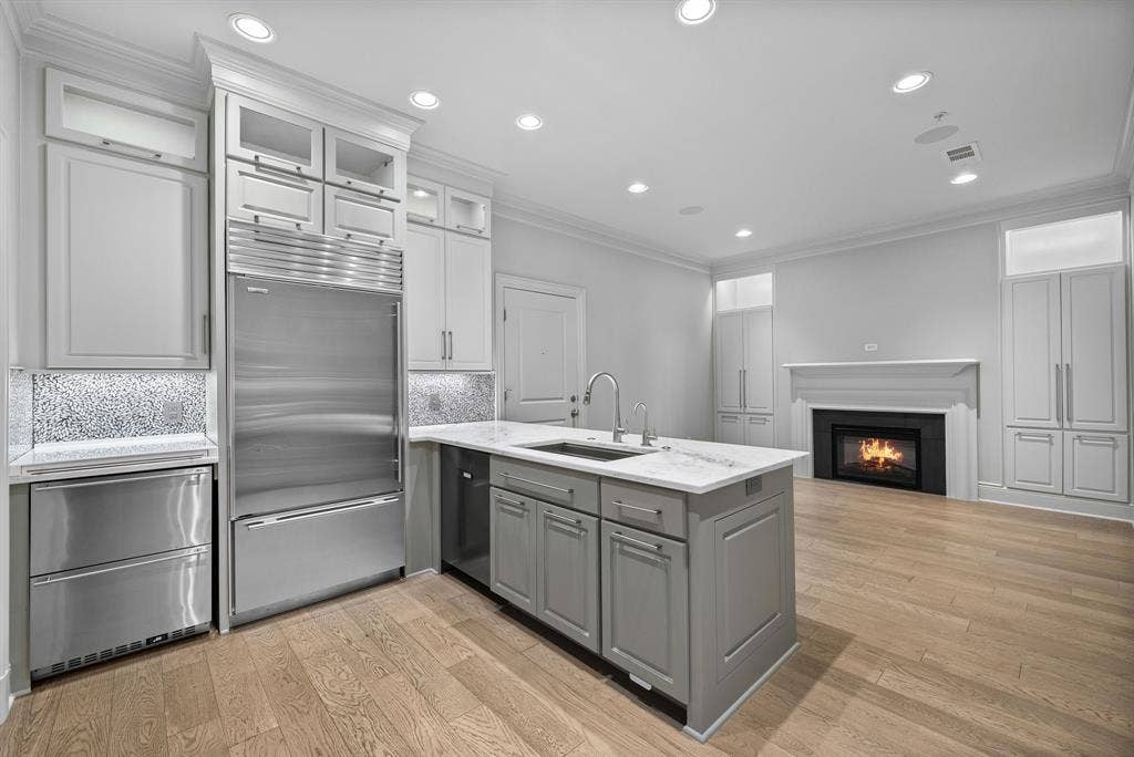 4240 Prescott Avenue, #8B, Dallas, TX 75219 Modern kitchen featuring a central island with an undermount sink, stainless steel appliances, light wood-finish flooring, recessed lighting, and a fireplace with a white mantel