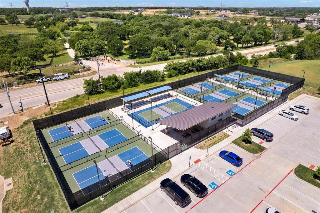 Dedicated pickleball facility featuring multiple courts, a central building with an expansive overhang, perimeter fencing, and an on-site parking area