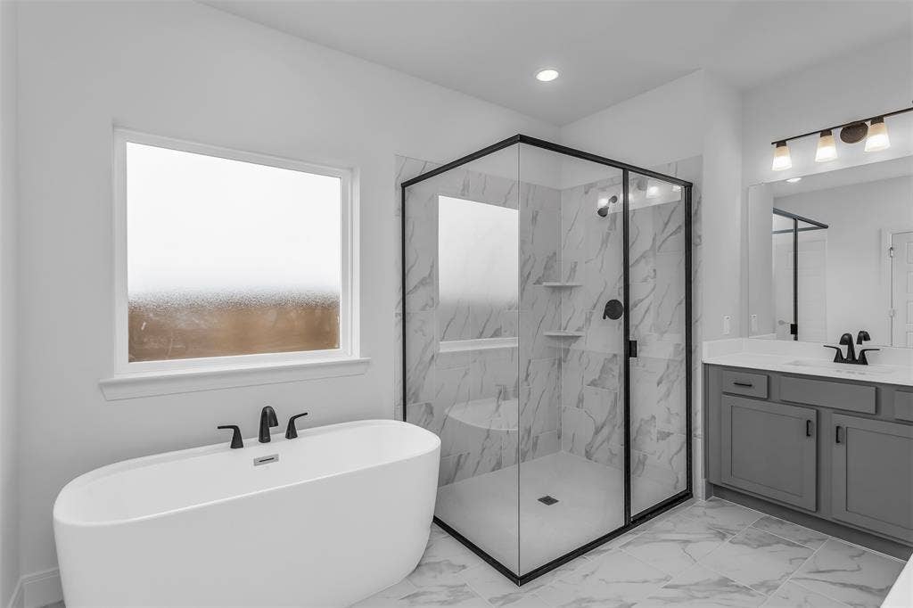 1031 Rylan Street, Midlothian, TX 76065 Modern bathroom featuring a freestanding soaking tub with matte black fixtures, a walk-in shower with black-framed glass enclosure and marble-look tile, a single vanity with grey cabinetry and white countertop, and large format marble-look floor tiling