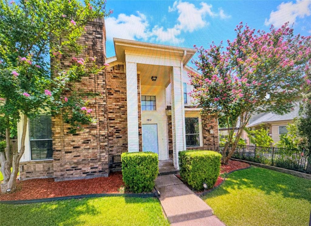 Brick facade residence with a columned portico entry, mature flowering trees, manicured hedges, and a concrete walkway