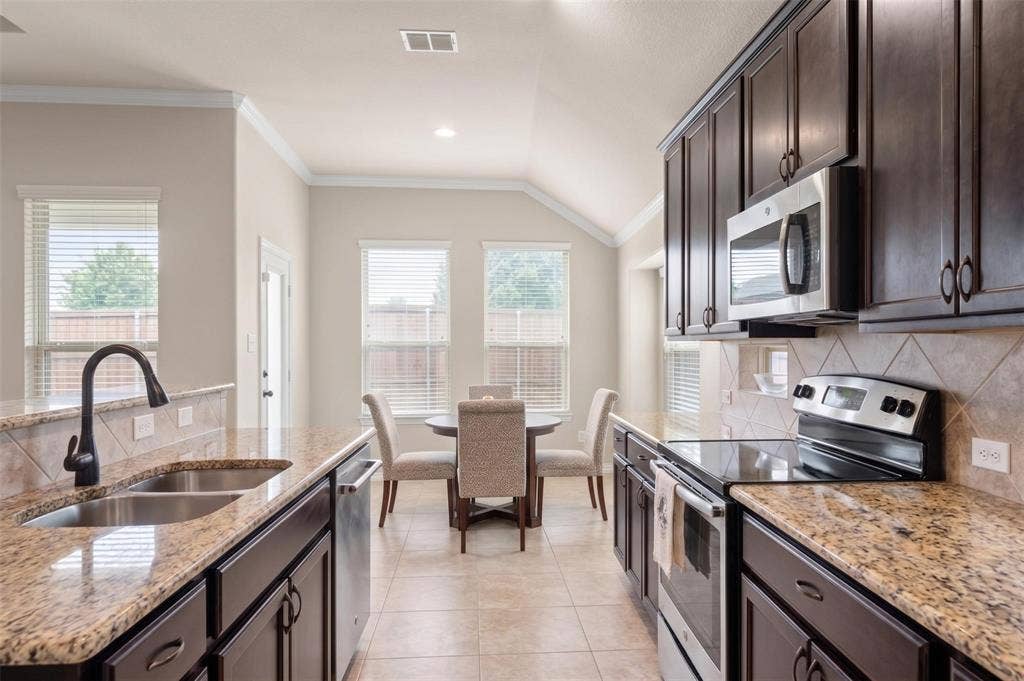 Galley kitchen featuring dark wood cabinetry, stainless steel appliances, granite countertops, and a tile backsplash