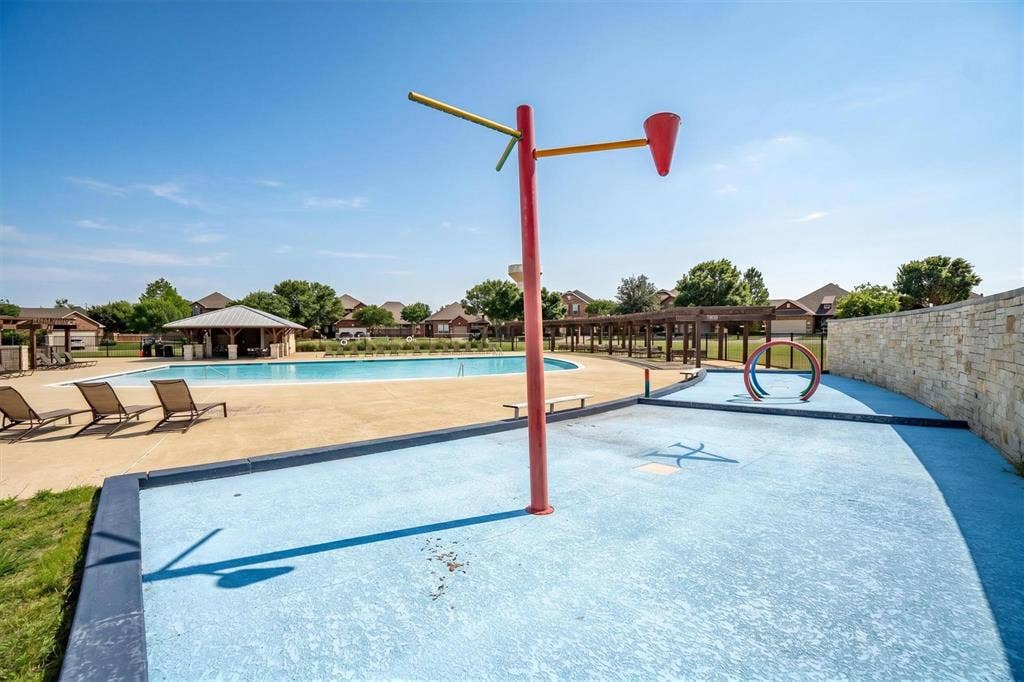 Outdoor aquatic area featuring a splash pad with water features, a swimming pool with a surrounding concrete deck, and a shaded pergola structure
