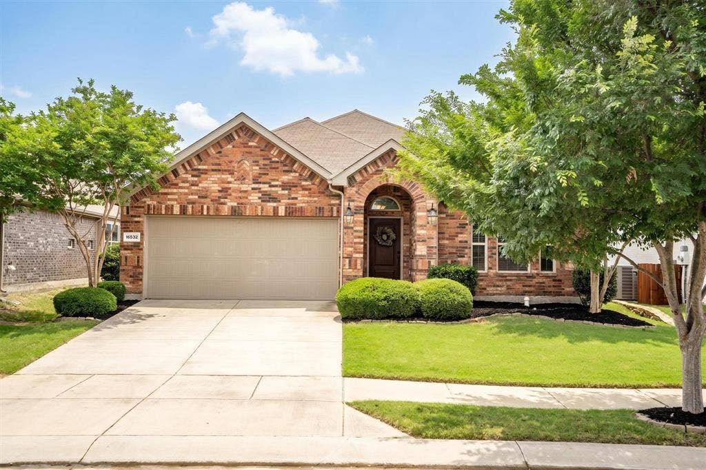 Brick facade residence featuring a two-car garage, arched entry with a dark wood door, mature trees, manicured lawn, and a concrete driveway