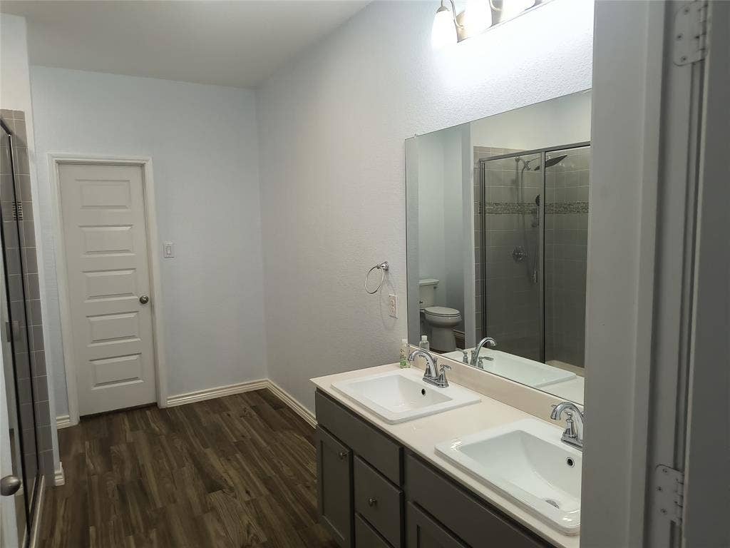 Double vanity with integrated sinks, wood-finish flooring, a frameless mirror, and a glass-enclosed shower with tiled walls