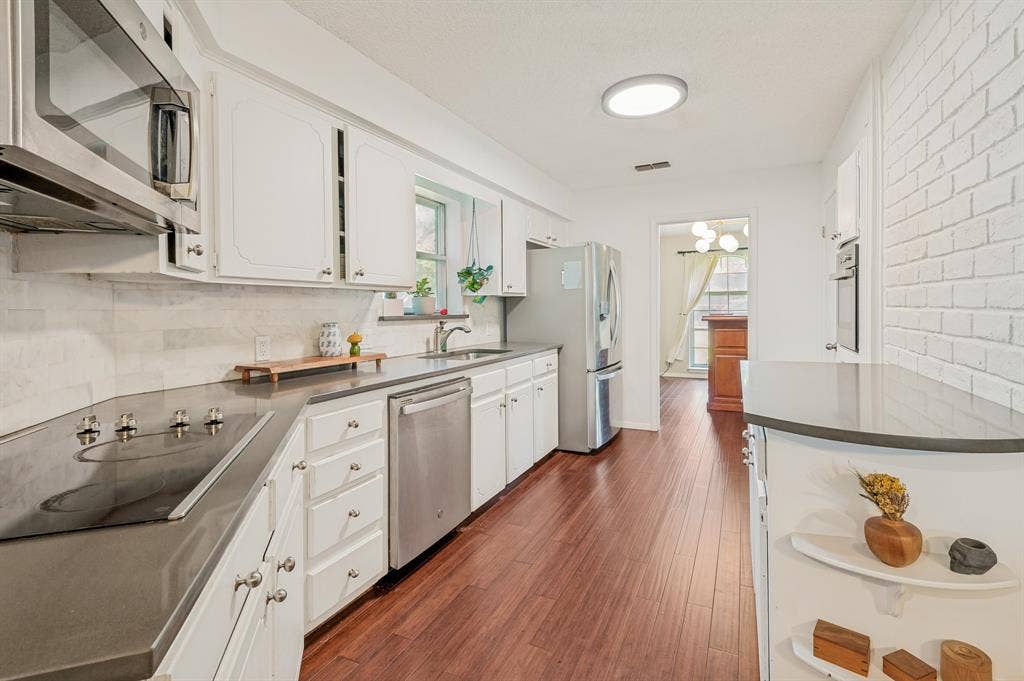404 Green River Trail, Fort Worth, TX 76103 Bright kitchen featuring white cabinetry, stainless steel appliances, dark countertops, wood-finish flooring, and a white brick-style accent wall