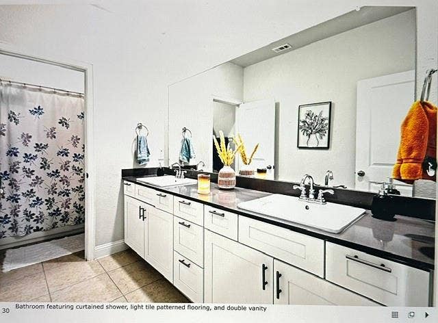 4220 Calla Drive, Forney, TX 75126 Bathroom featuring a double vanity, dark countertop, dual chrome faucets, white cabinetry with dark hardware, and light-toned tile flooring
