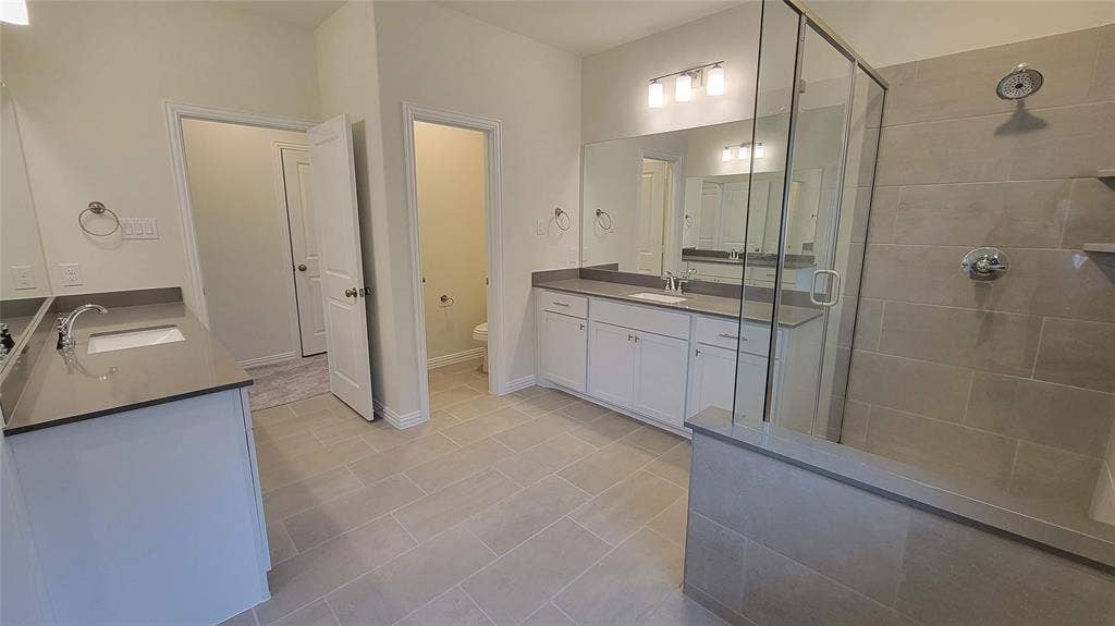 11346 Sugarlands Drive, Frisco, TX 75035 Spacious bathroom featuring dual vanities with integrated sinks and dark countertops, a glass-enclosed shower with tile surround, and light-toned tile flooring