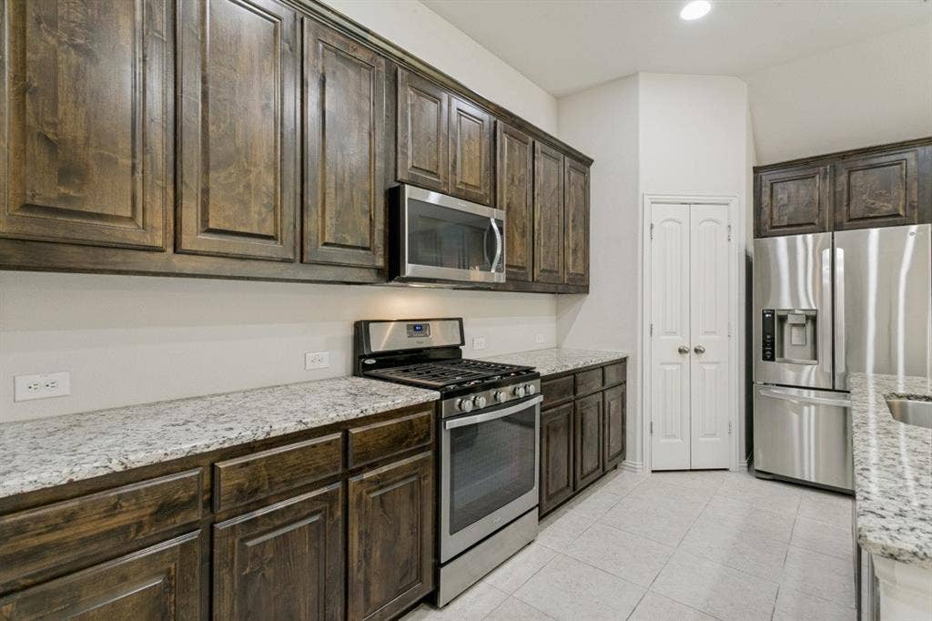 Kitchen featuring dark wood cabinetry, light speckled countertops, stainless steel appliances, and white tile flooring