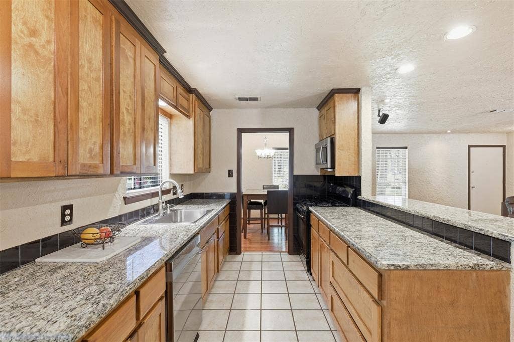 3702 Hastings Drive, Arlington, TX 76013 Galley-style kitchen with granite countertops, wood cabinetry, double sink, tile flooring, and multiple windows.