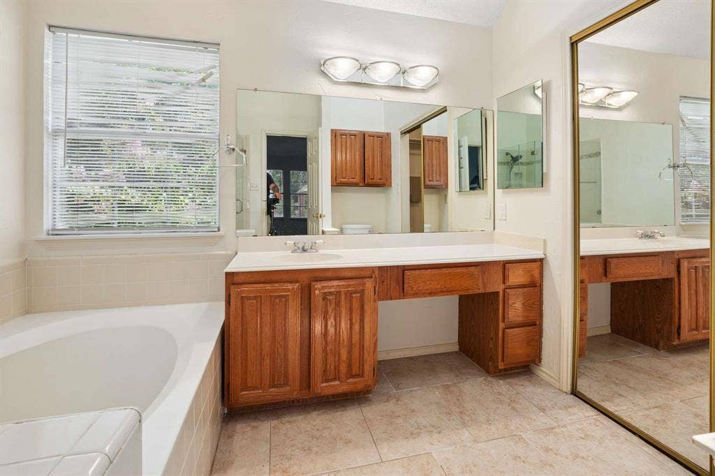 Bathroom featuring a built-in soaking tub, wood-finish vanity with countertop, wall-mounted mirrors, vanity lighting, and tile flooring