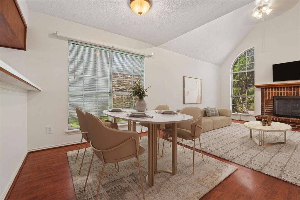 Open-concept living area with vaulted ceilings, a brick-front fireplace, wood-finish flooring, and a large arched window