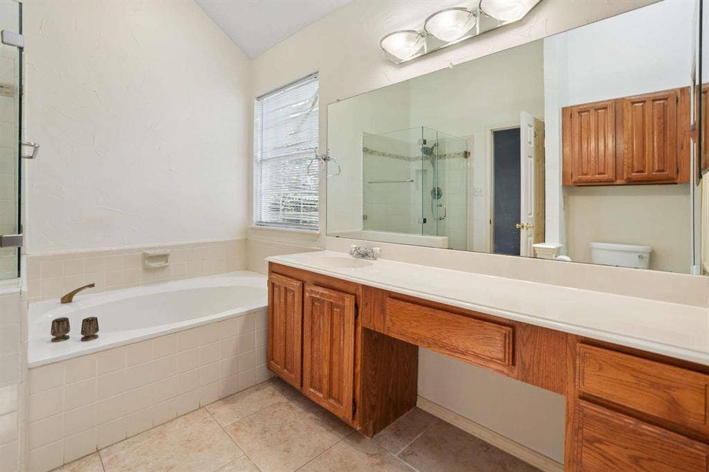Bathroom featuring a built-in bathtub with tiled surround, a separate glass-enclosed shower, wood-finish vanity cabinetry with an integrated countertop, a full-width mirror, and tile flooring