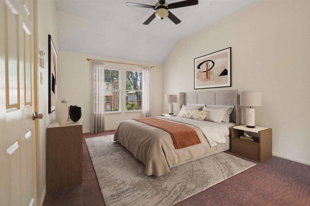 Vaulted ceiling bedroom featuring plush carpeting, a ceiling fan with light fixture, and two large window panes with sheer white curtains
